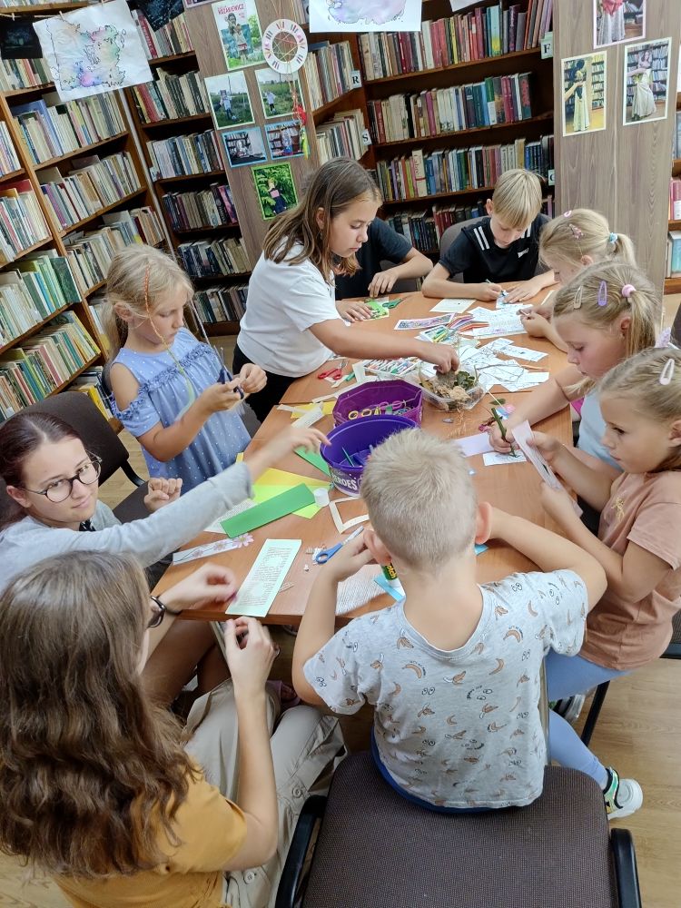 Grupa siedzących i stojących przy stole dzieci wykonujących prace plastyczne. W tle regały z książkami.