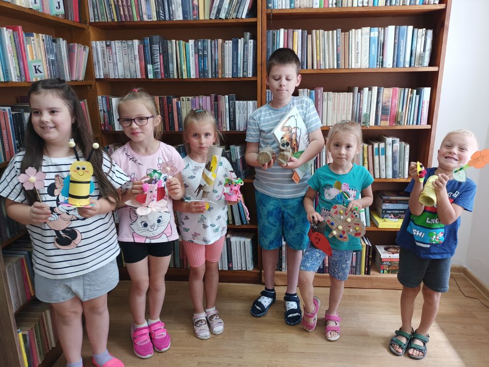 Sześcioro stojących dzieci trzymających w rękach papierowe owady. W tle regały z książkami.
