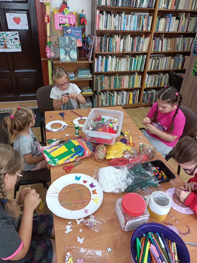 Pięć dziewczynek siedzących wokół stołu wykonujących prace plastyczne. W tle, po prawej, regały z książkami, z lewej drzwi