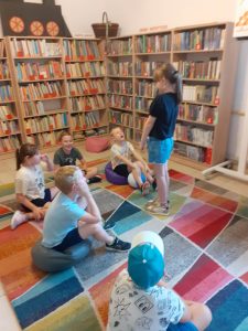 Na kolorowym dywanie siedzi grupka dzieci. Przed nimi stoi dziewczynka. W tle regały z książkami.