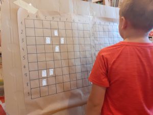 Na zdjęciu chłopczyk stoi przed dużą planszą na, której narysowane są kwadraty i przyklejone karteczki ze śladami stóp.