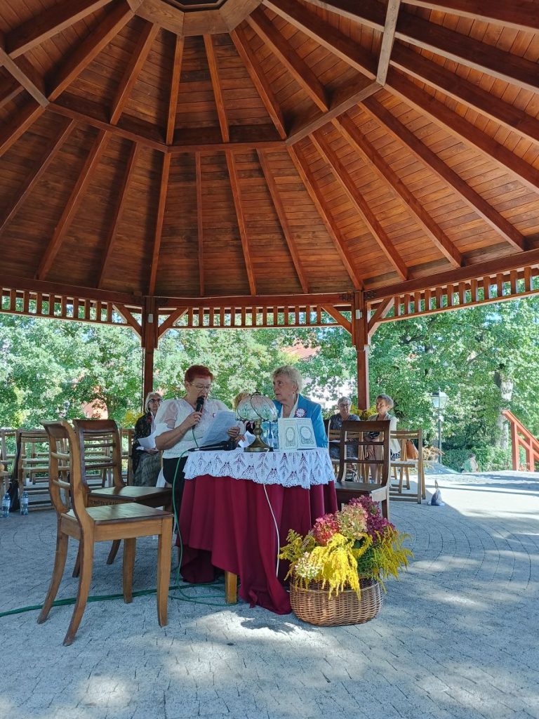 Siedzące przy stoliku dwie kobiety. Kobieta z lewej czyta. W Tle grupa siedzących przodem kilku osób. Za nimi drzewa. Nad nimi widok zadaszenia dużej altany.