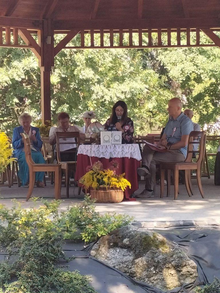 Od lewej siedząca i czytająca przy stoliku kobieta, obok niej mężczyzna. W Tle grupa siedzących przodem kilku osób. Za nimi drzewa. Nad nimi widok zadaszenia dużej altany.