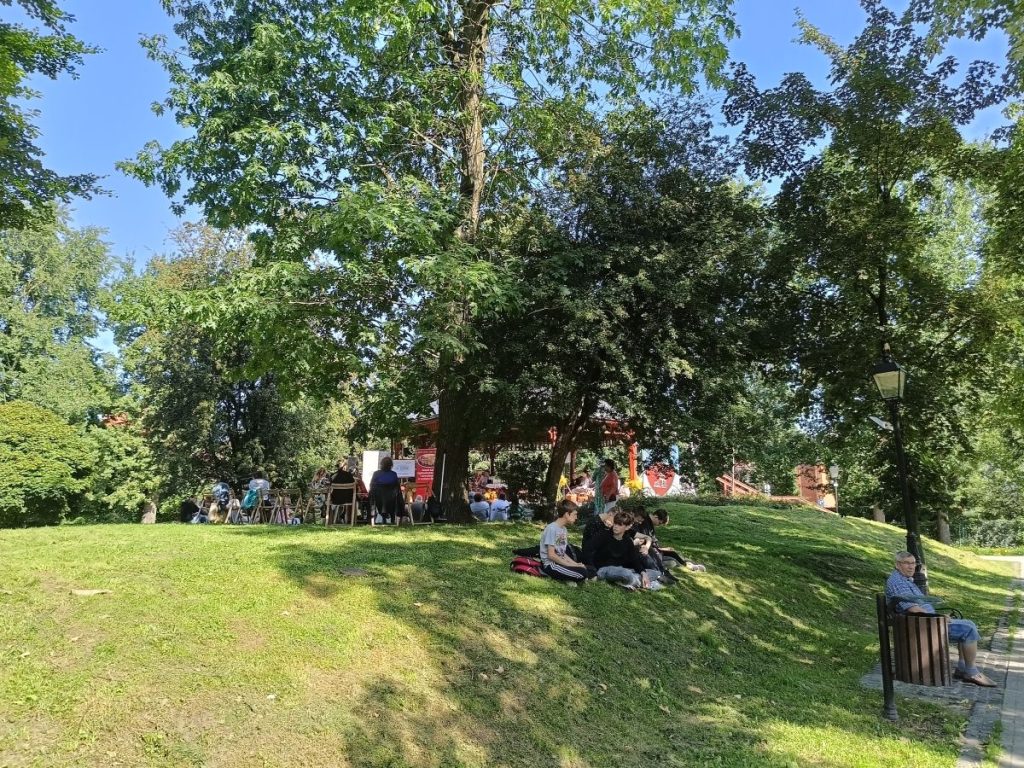 Na pierwszym planie grupa kilku osób siedzących na trawie. Za nimi drzewa, pomiędzy drzewami grupa siedzących tyłem osób.