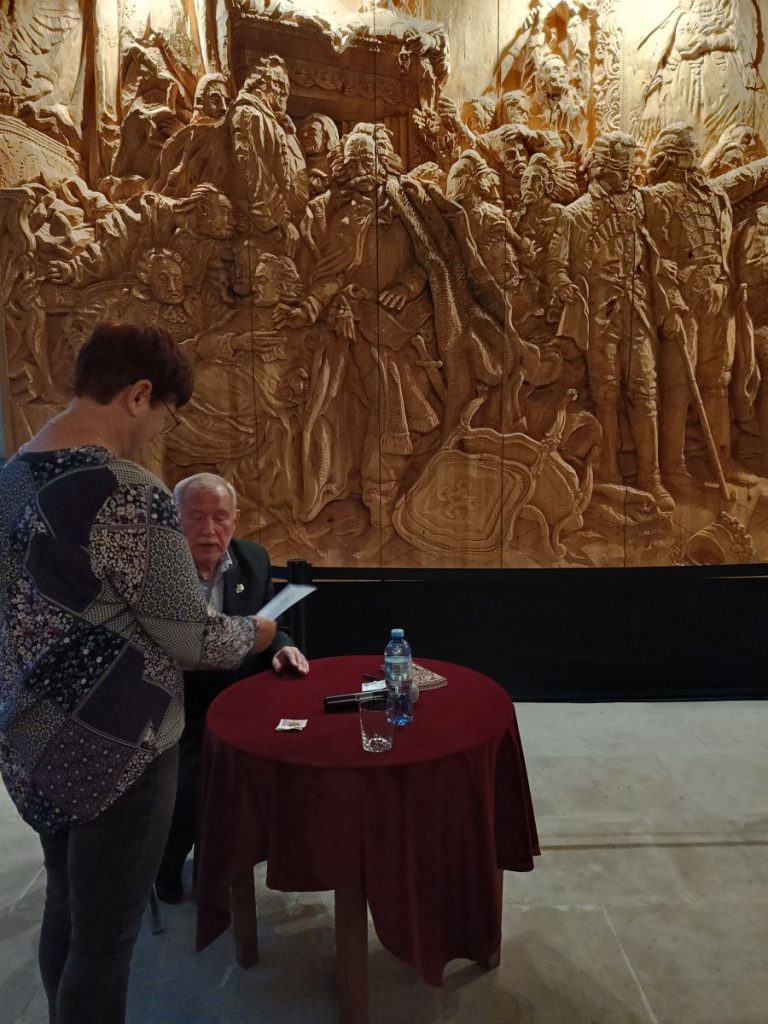 Od lewej stojąca tyłem kobieta obok siedzącego przy stole mężczyzny zwróconego przodem. W tle widok monumentalnej rzeźby.