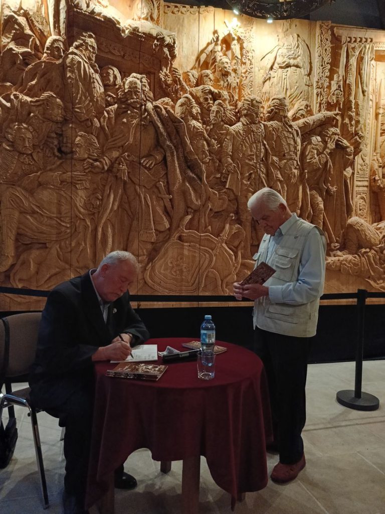 Po lewej siedzący przy stoliku mężczyzna podpisujący książkę. Po prawej stojący bokiem mężczyzna trzymający w rękach kartkę. W tle widok monumentalnej płaskorzeźby.