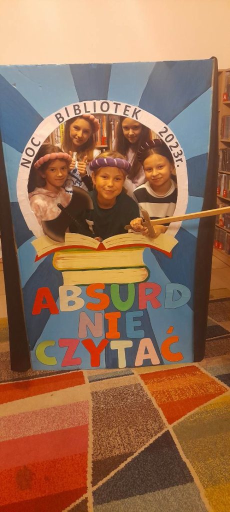Dziewczynki w toczkach na głowie stoją w okienku. Fotobudka wykonana z dużego kartonu w kolorze niebieskim, z napisami ‘’Noc bibliotek 2023 rok’’ , ‘’Absurd nie czytać’’, na którym namalowane są trzy książki.