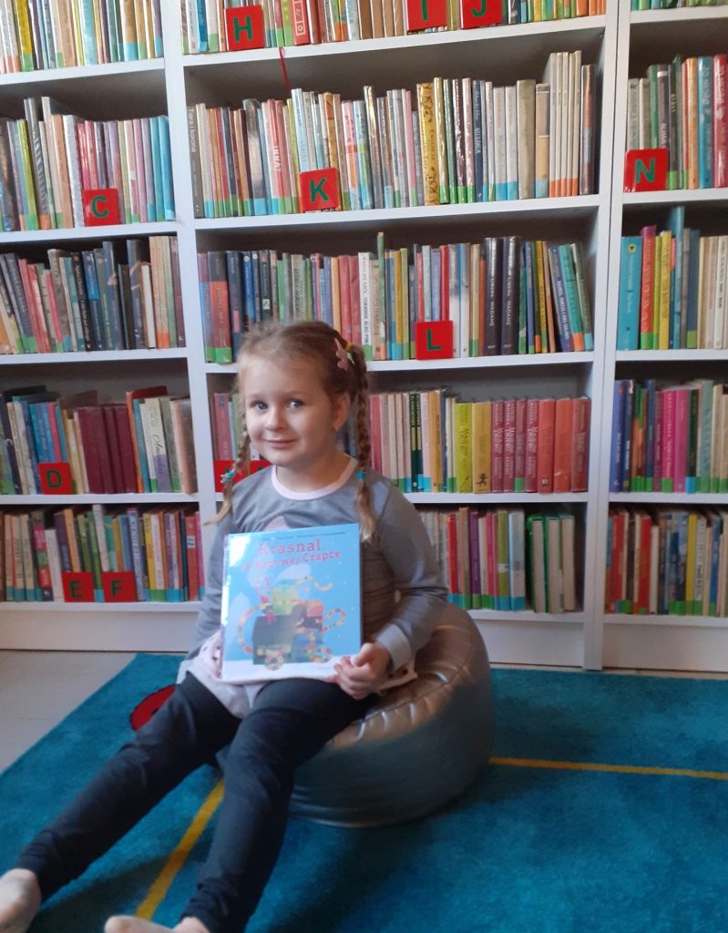 Na dywanie, na srebrnej pufie siedzi dziewczynka. W rękach trzyma książkę. W tle regały z kolorowymi książkami.