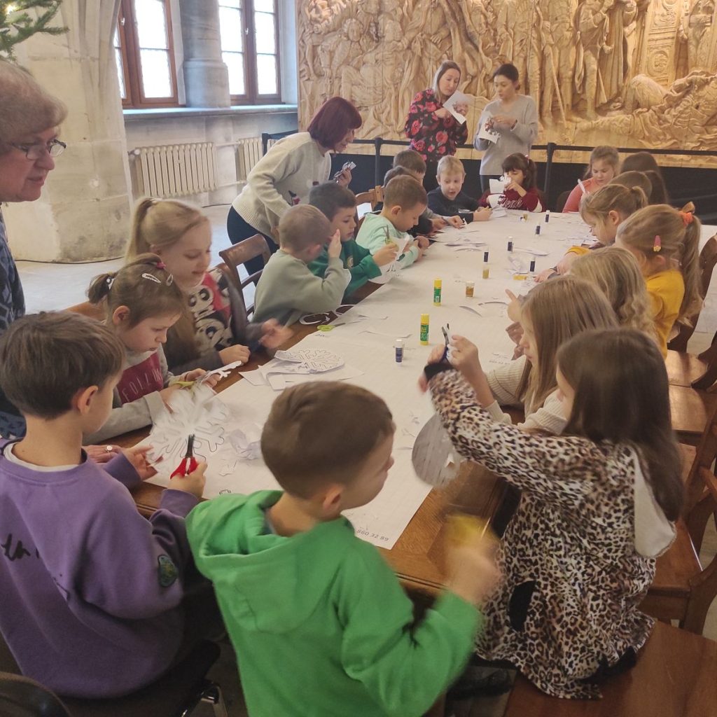 Wzdłuż stołu, który znajduje się na środku zdjęcia siedzą dzieci. Dzieci trzymają w rękach nożyczki. Z lewej strony nad dziećmi nachyla się jedna z pań. Na końcu stołu stoją dwie panie, w rękach trzymają kartki.
