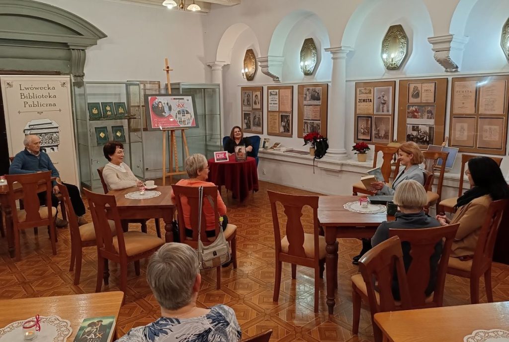 2. Grupa osób siedzących tyłem przy stolikach. Naprzeciw siedząca przy stoliku kobieta. W tle, od lewej, rollup z napisem Lwówecka Biblioteka Publiczna, szklane gabloty, plansze z fotografiami, a nad nimi kinkiety.