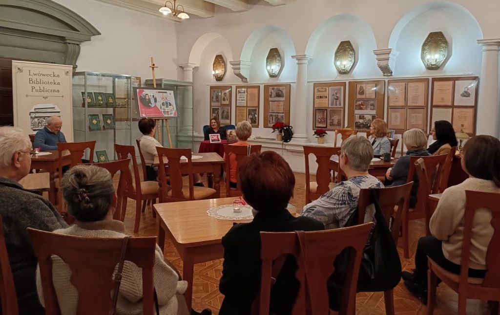 4. Grupa osób siedzących tyłem przy stolikach. Naprzeciw siedząca przy stoliku kobieta. W tle, od lewej, rollup z napisem Lwówecka Biblioteka Publiczna, szklane gabloty, plansze z fotografiami, a nad nimi kinkiety.