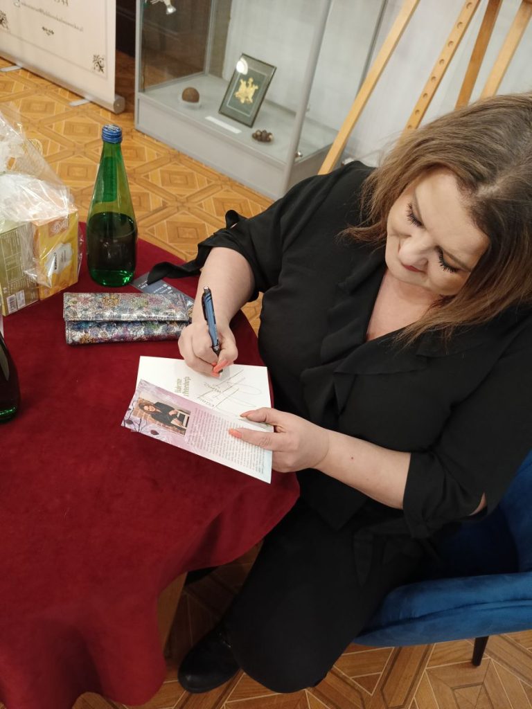 7. Siedząca przy stoliku kobieta podpisująca książkę. Obok niej, na stoliku, portfel oraz szklana butelka.