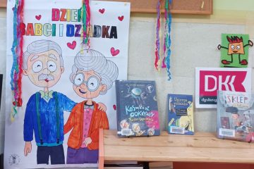 1. Z lewej strony znajduje się duży, kolorowy plakat z postaciami Babci i Dziadka. Z prawej strony na półce stoją książki, a nad nimi wisi kilka kolorowych bibułek.