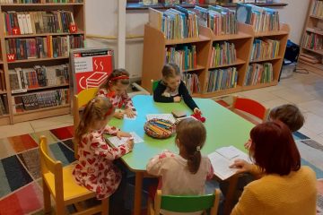 Dzieci siedzą przy stoliku, na którym leżą kredki, malowanki, włóczka, tekturowe serca. Pani kuca między dwoma dziewczynkami, a przed nią na stoliku leży otwarta książka. W tle duże okna i regały z kolorowymi książkami.