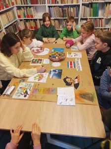 Dzieci siedzą wkoło stołu, na którym leżą: koszyk z kredkami, kleje oraz szary papier pokolorowany i ozdobiony kolorowymi kartkami. W tle regały z książkami.