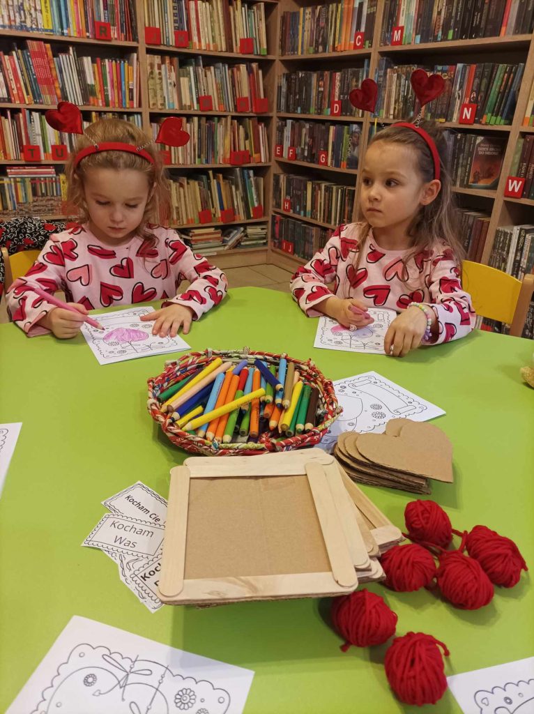 Przy zielonym stoliku siedzą dwie dziewczynki, które kolorują obrazek. Przed nimi leżą: kredki, czerwone włóczki, serca i ramki tekturowe. W tle regały z książkami.