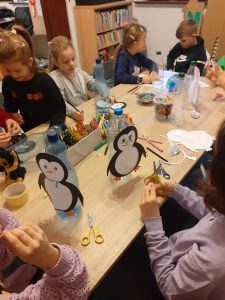 Na stole leżą: kredki, butelki plastikowe z przyklejoną postacią pingwina. Dzieci siedzą wkoło stołu, a w tle są regały z książkami.
