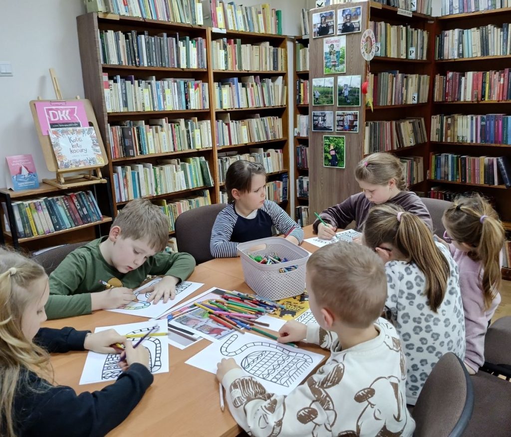 Grupa dzieci siedząca przy stole, malująca obrazki. Na stole kartki i kredki. W tle, od lewej, mały regał z książkami, następnie widok dużych regałów z książkami.