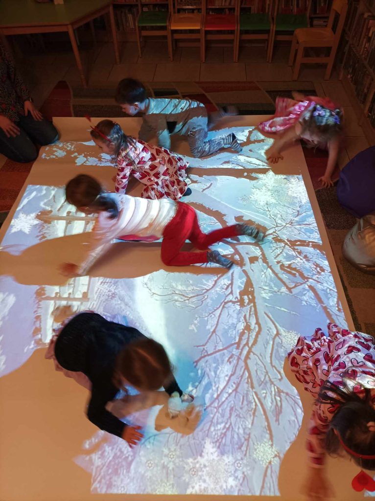 Na podłodze leży mata – podłoga interaktywna, na której bawią się dzieci. W tle krzesełka i stolik.
