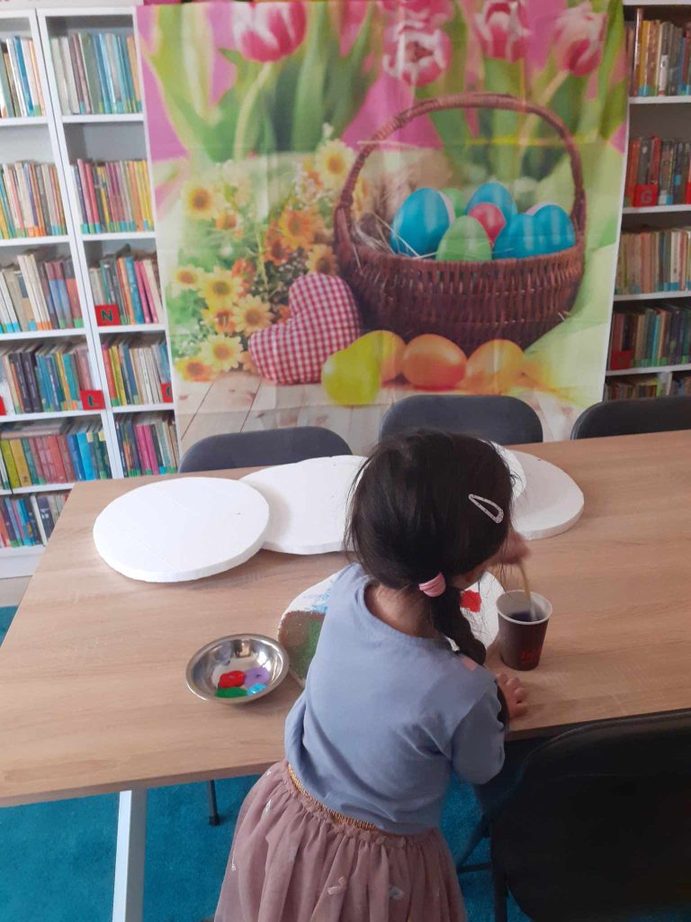Dziewczynka siedzi przy stoliku, na którym leżą styropianowe jajka, farby i kubek z wodą. W tle kolorowy, świąteczny obrazek i regały z książkami.