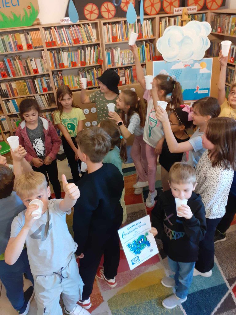 Grupka dzieci trzyma kubeczki z wodą. W tle plansza z plakatem i chmurą oraz regały z książkami.