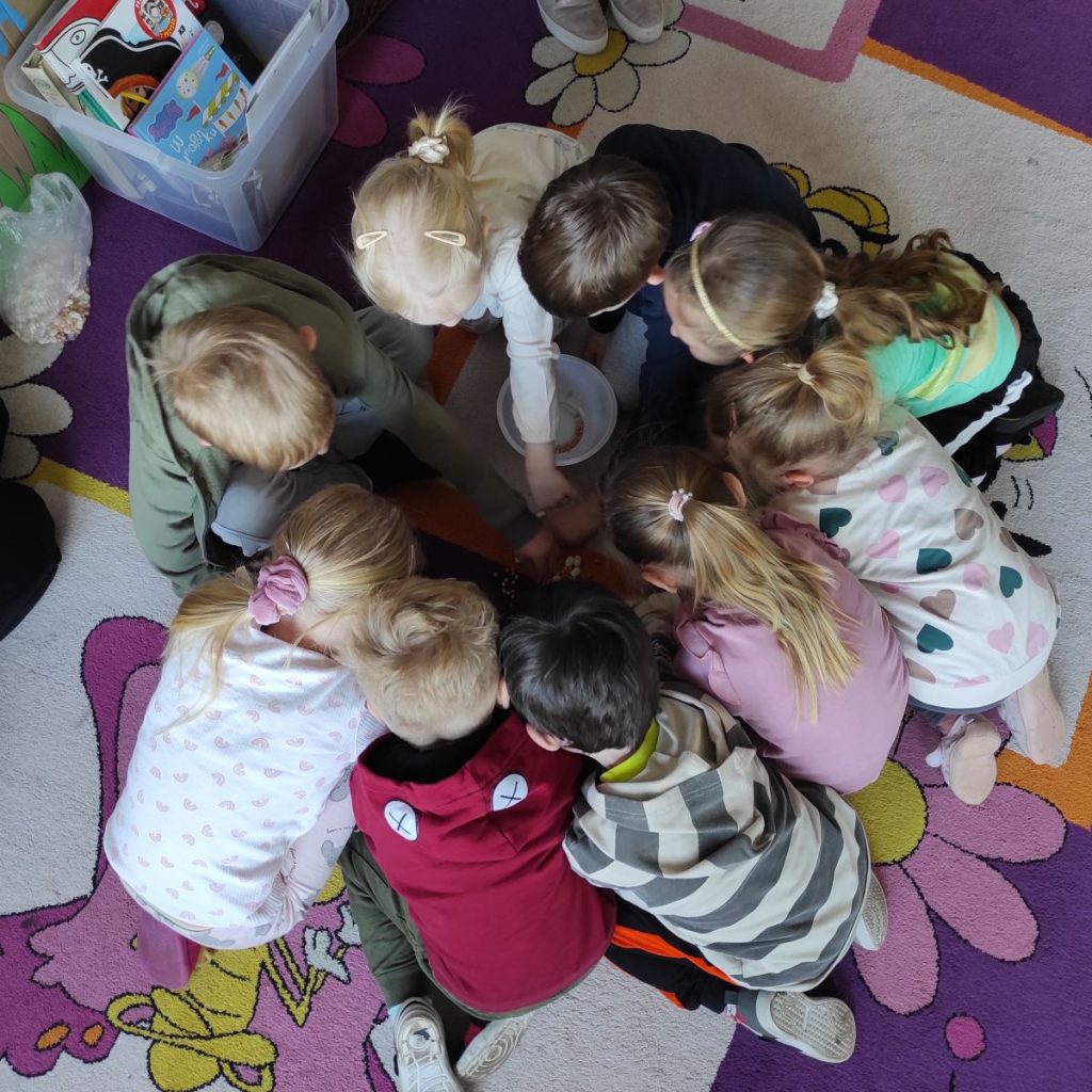 Na kolorowym dywanie siedzą w kole dzieci, a w nim miska z fasolą i grochem. Obok stoi pudełko z książkami i woreczek z fasolą i grochem.