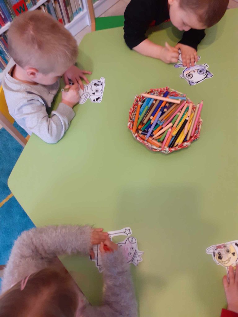Przy stoliku siedzą dzieci, kolorują postacie z bajek. Na środku stołu leży koszyk z kolorowymi kredkami.