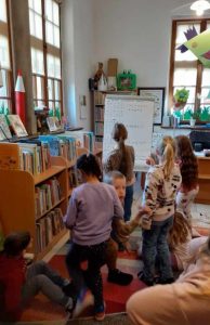 Grupka dzieci stoi przed planszą z zadaniem. W tle regały z książkami, biurko pani bibliotekarki, okna.