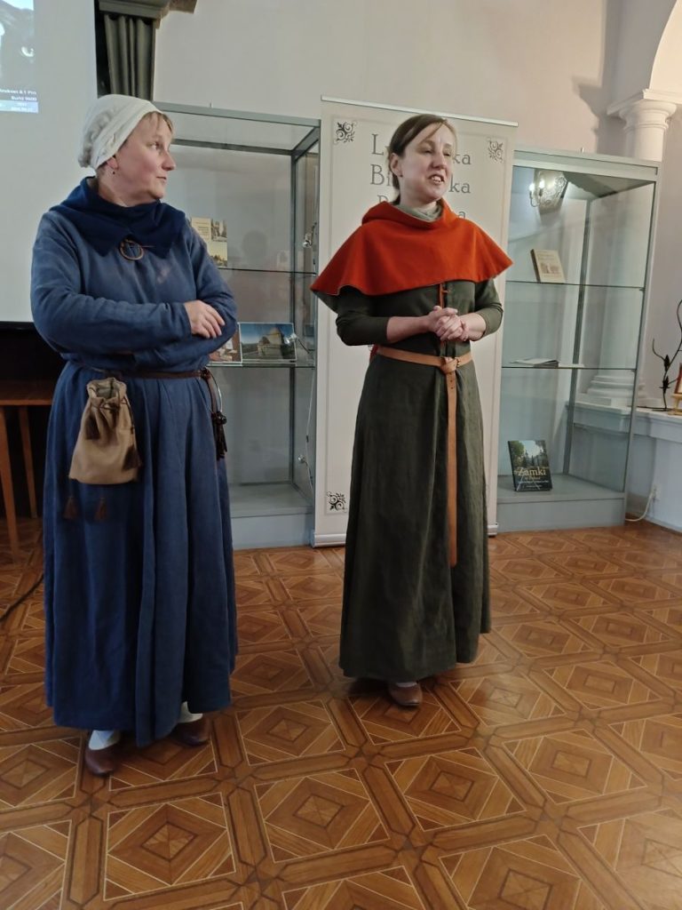 Dwie stojące kobiety, ubrane w długie szaty. Kobieta z lewej z czepkiem na głowie. Za nimi fragmenty dwóch szklanych gablot z książkami wewnątrz.